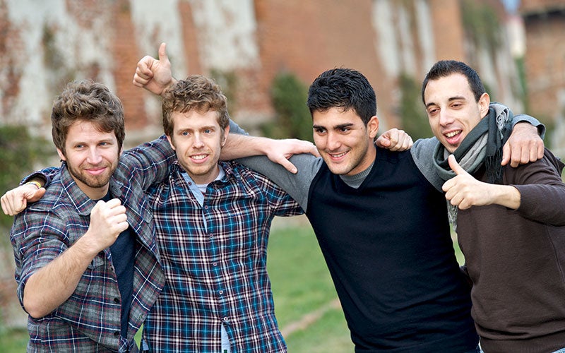 Four male students stand with the arms around each other's shoulders in the grass on campus.